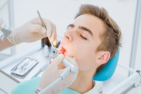 The image shows a dental professional performing a dental procedure on a patient, with the patient seated in an examination chair and the professional working at the dental station.