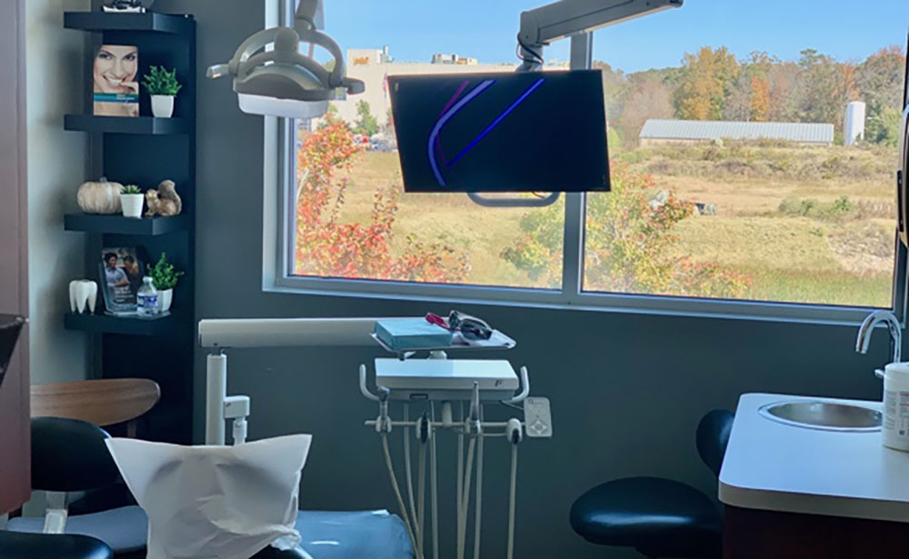 The image shows an interior view of a dental practice with various pieces of equipment and a large window showing outdoor scenery.