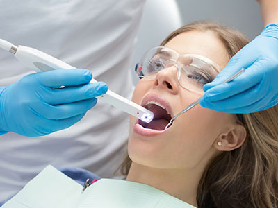 The image shows a dental professional performing a teeth whitening procedure on a seated patient, with the dentist holding a device to the patient s mouth and the patient wearing protective eyewear.